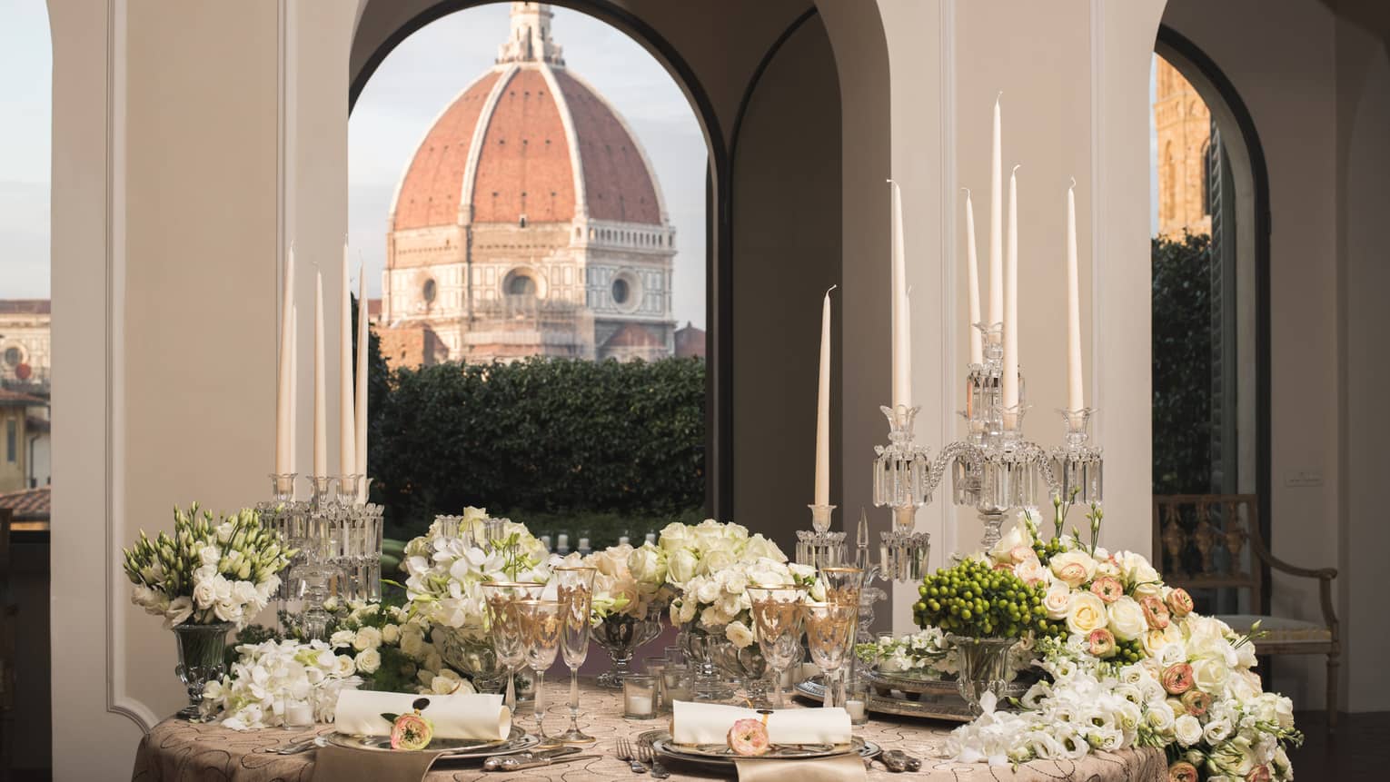 Elegant dining table set with candles, flowers, and glassware, with a large dome visible through arched windows in the background.
